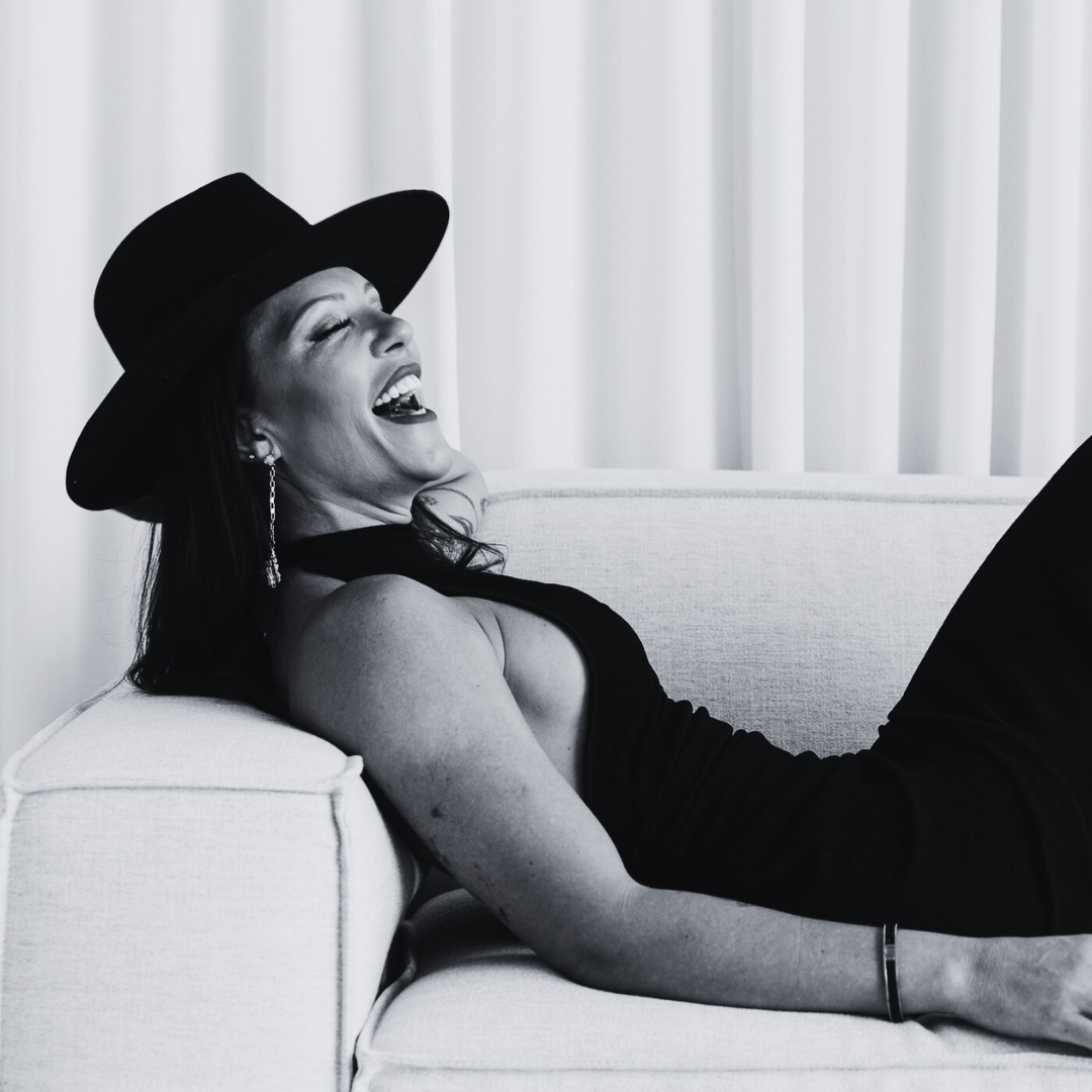 Mariana laughing as she relaxes on a sofa, wearing a black hat