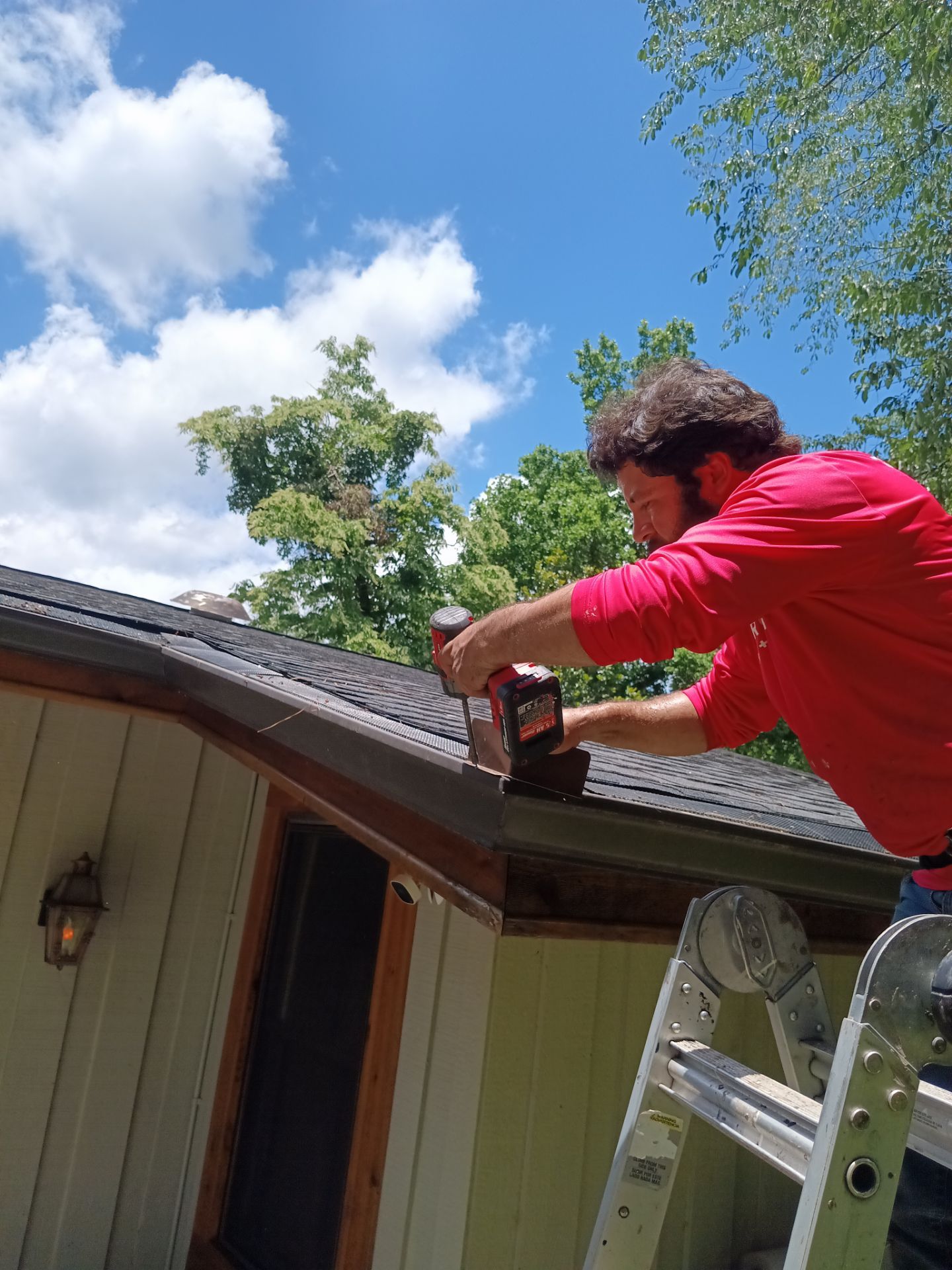 Shelter Guard crew member in a red shirt using a drill on a gutter from a ladder