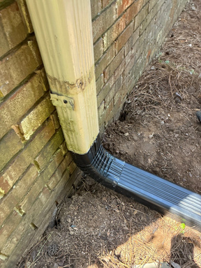 Black downspout elbow at the base of a wood post against a brick wall