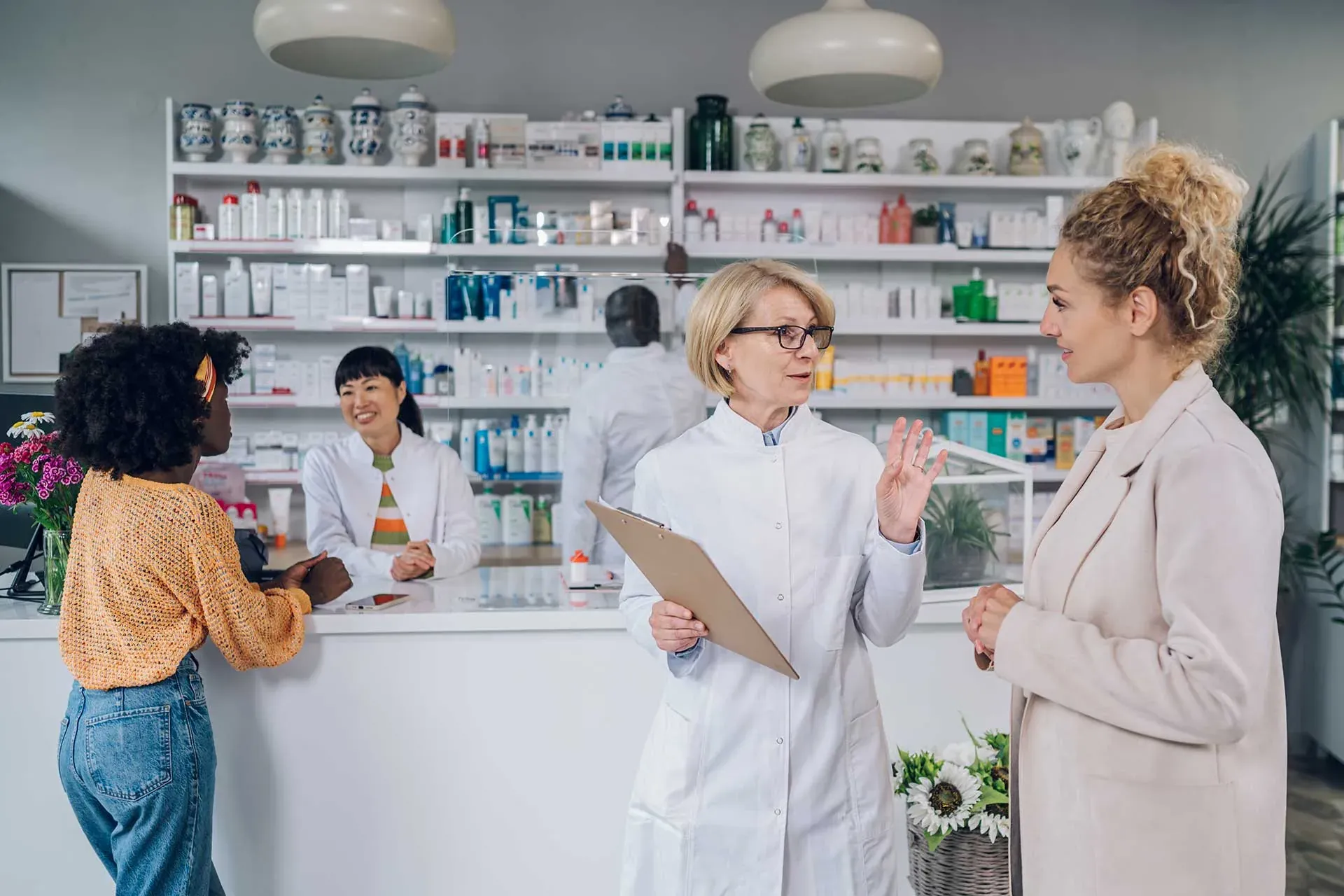 Two pharmacists talk with customers across the counter of a bright community pharmacy