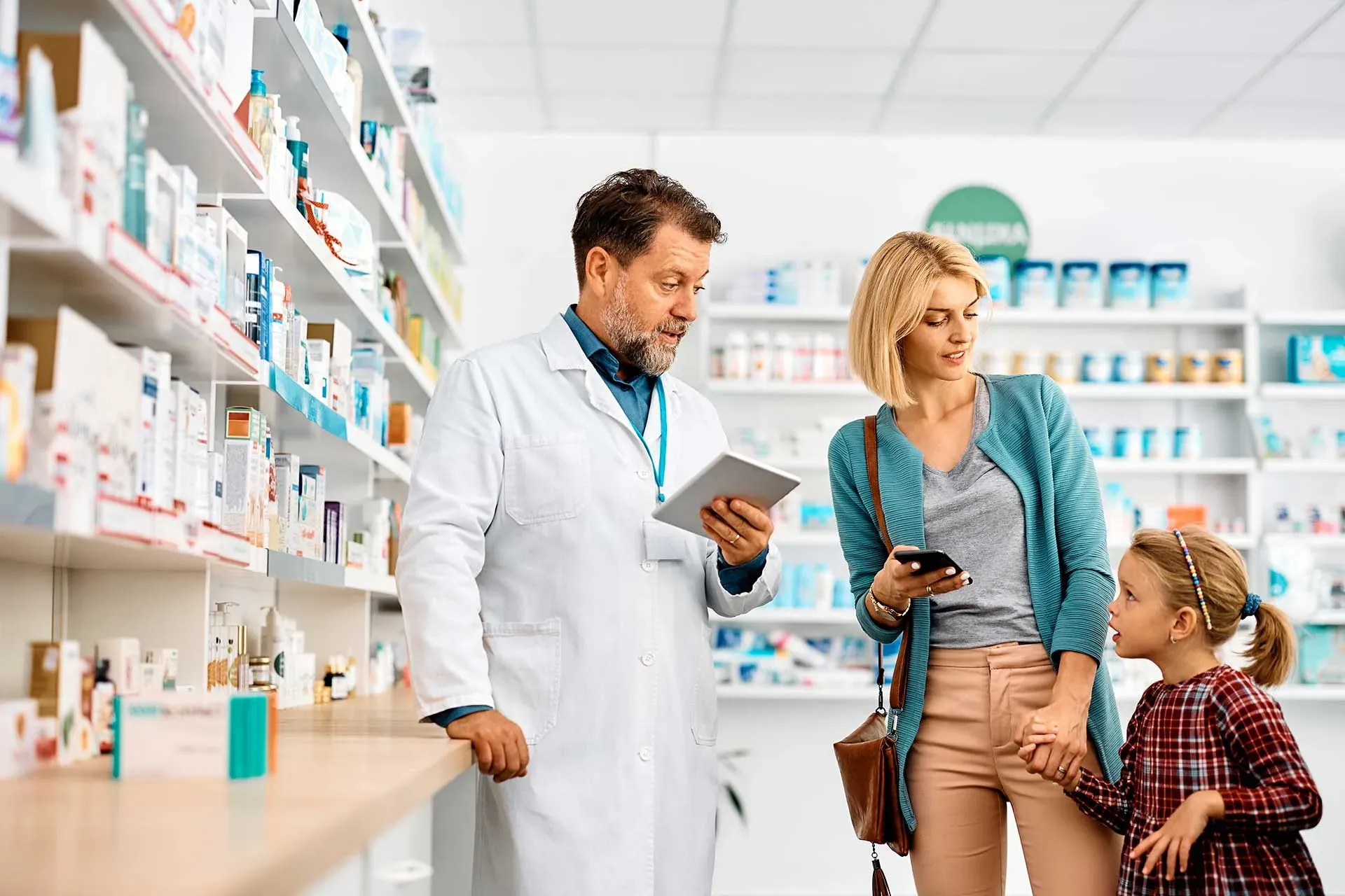 A pharmacist with a tablet helps a mother and daughter in a community pharmacy aisle
