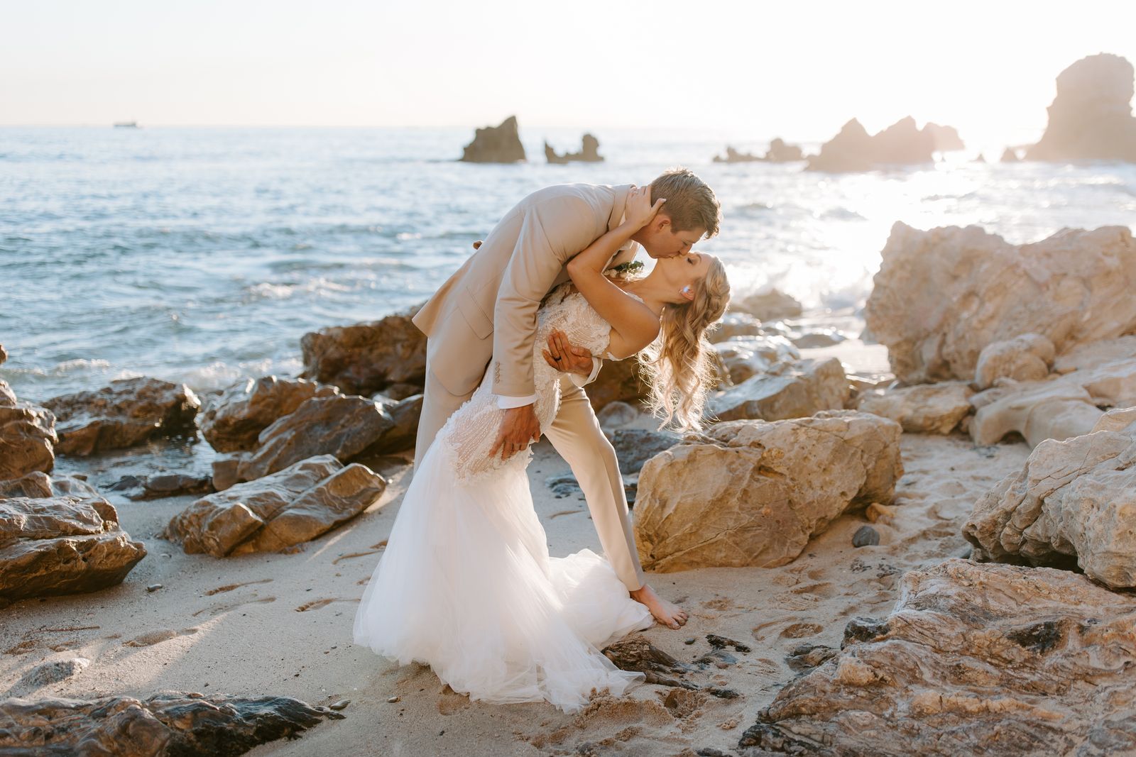 A groom dipping his bride into a kiss on the rocks by the ocean
