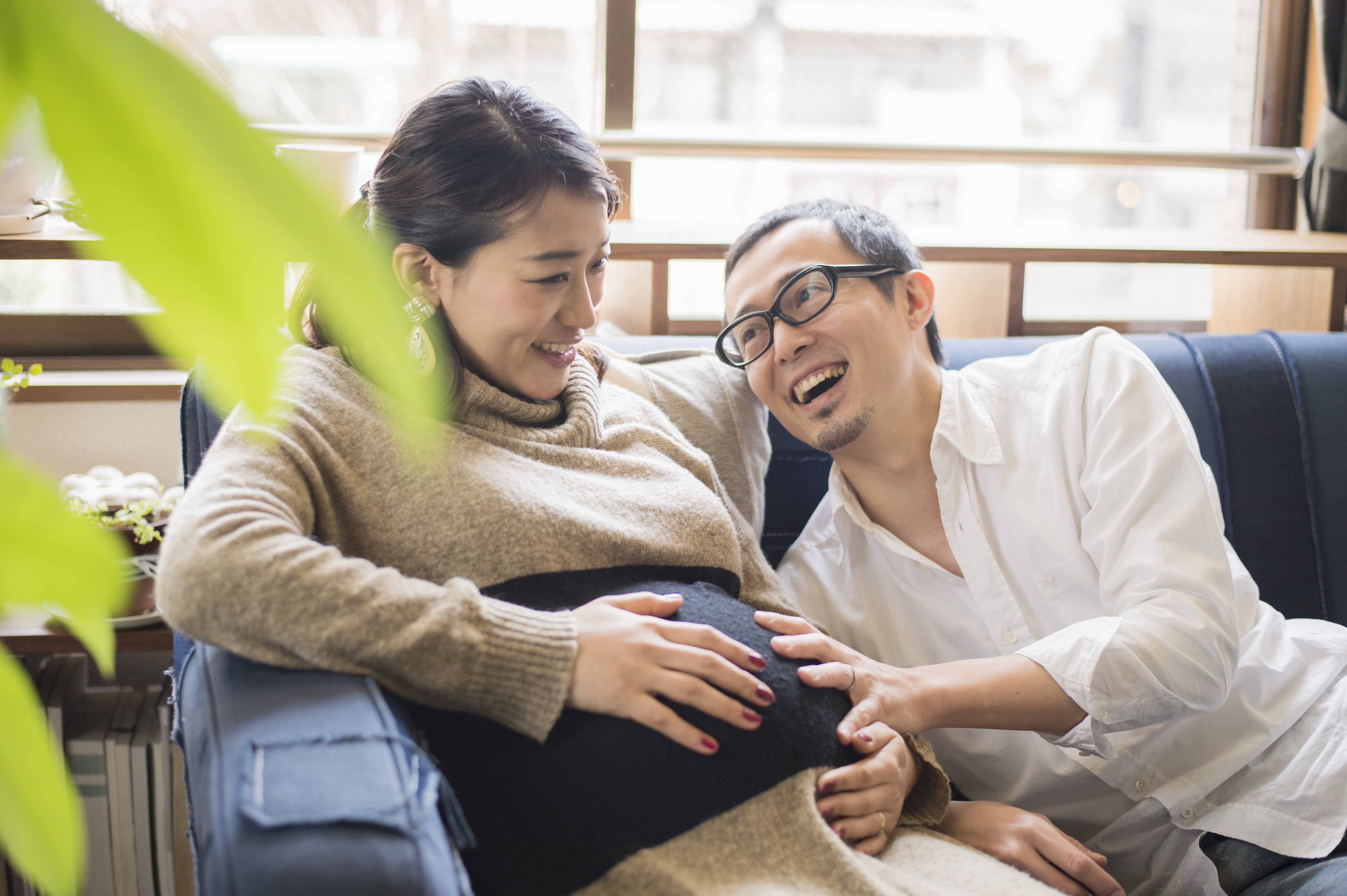 A pregnant woman sits on a couch while a man beside her smiles and gently touches her belly. They appear to be in a living room with natural light from a window.