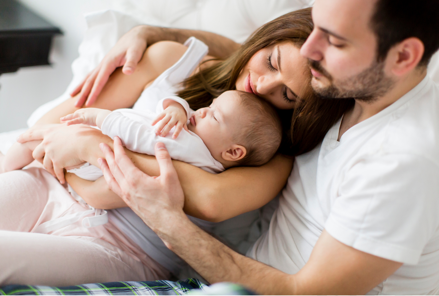 A man and woman gently hold their baby while lying together on a bed, showcasing a moment of family intimacy.