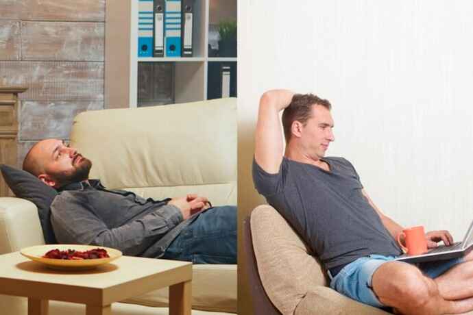 Man relaxing on couch, one lying down and another seated with a laptop, both at ease.