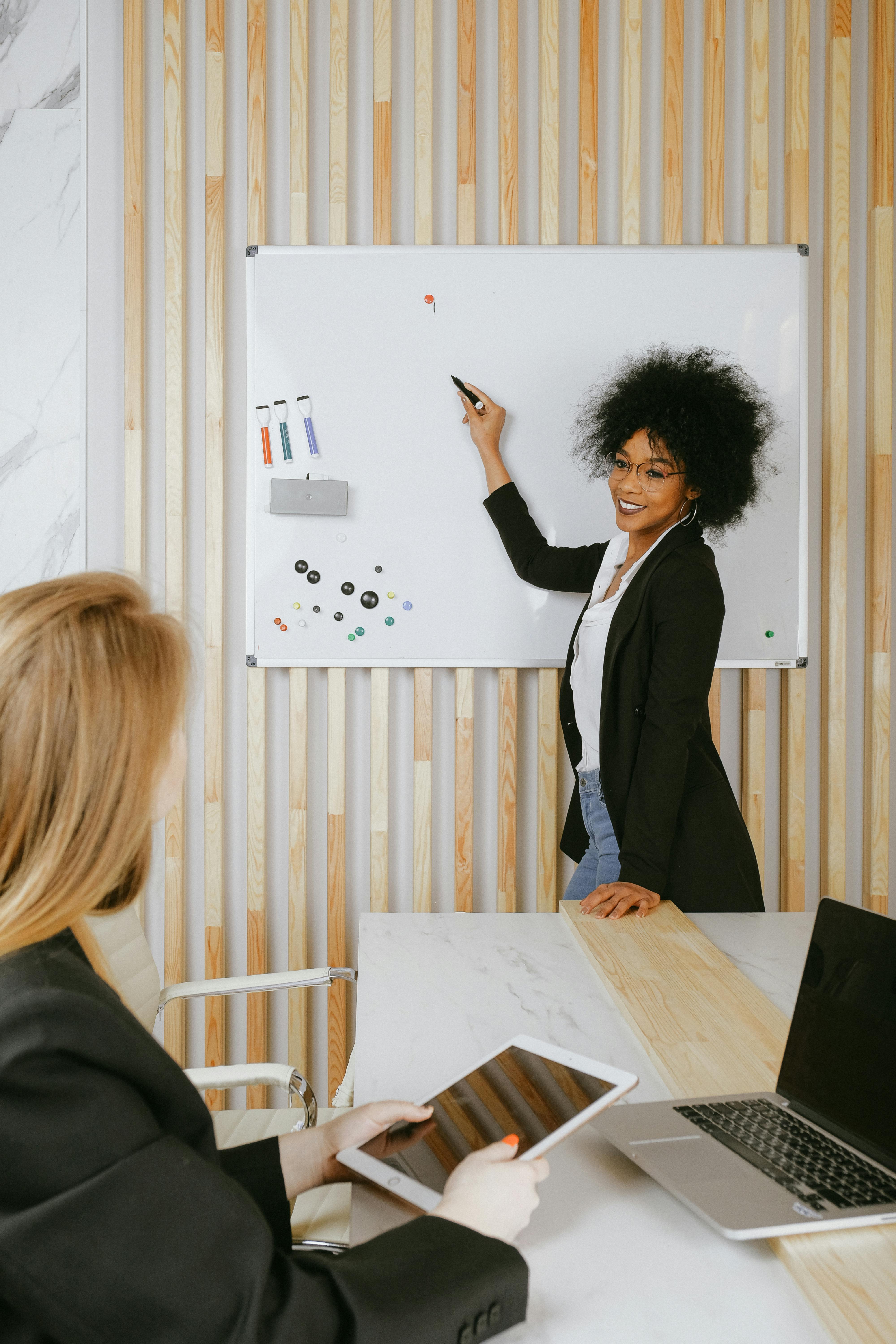 A picture of a woman writing on a whiteboard and looking back at a woman seated at a table behind her. 