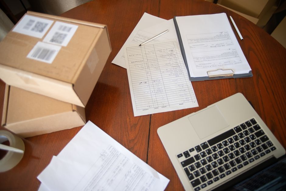 Wooden desk with a clipboard of forms, loose paperwork, pencils, tape and small cardboard boxes beside an open laptop.