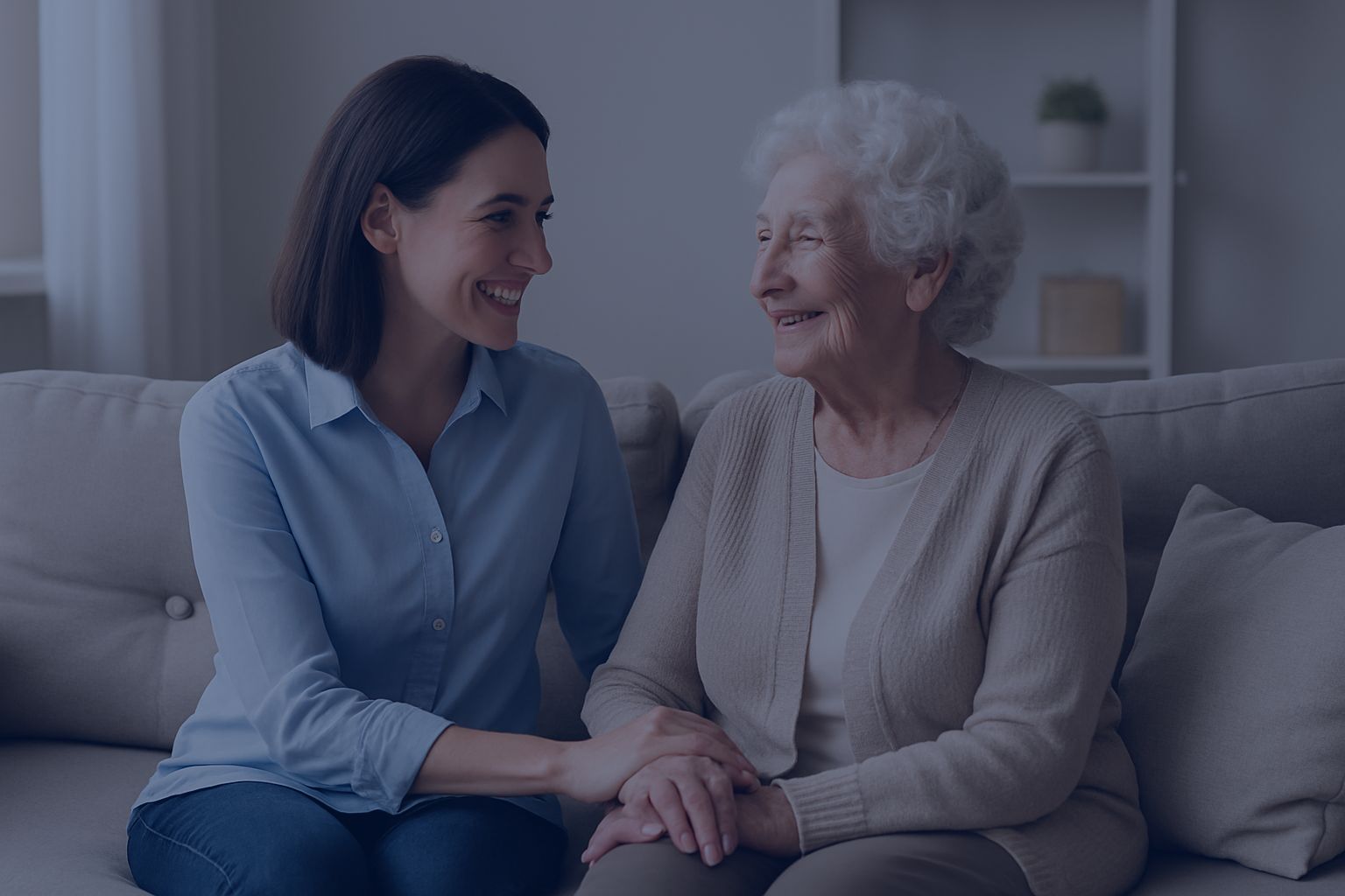 A caregiver and an older woman smiling together on a sofa, holding hands
