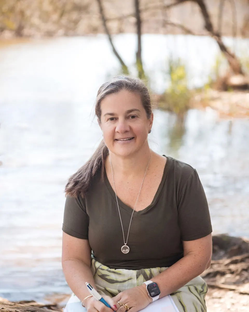 Tammy Schwarzl smiling, seated with a notebook beside a calm Central Coast lagoon