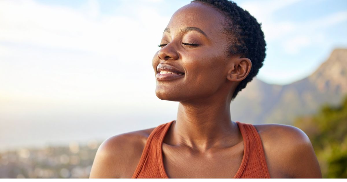 Midlife woman with eyes closed breathing deeply outdoors in sunlight, symbolizing staying grounded during uncertain times