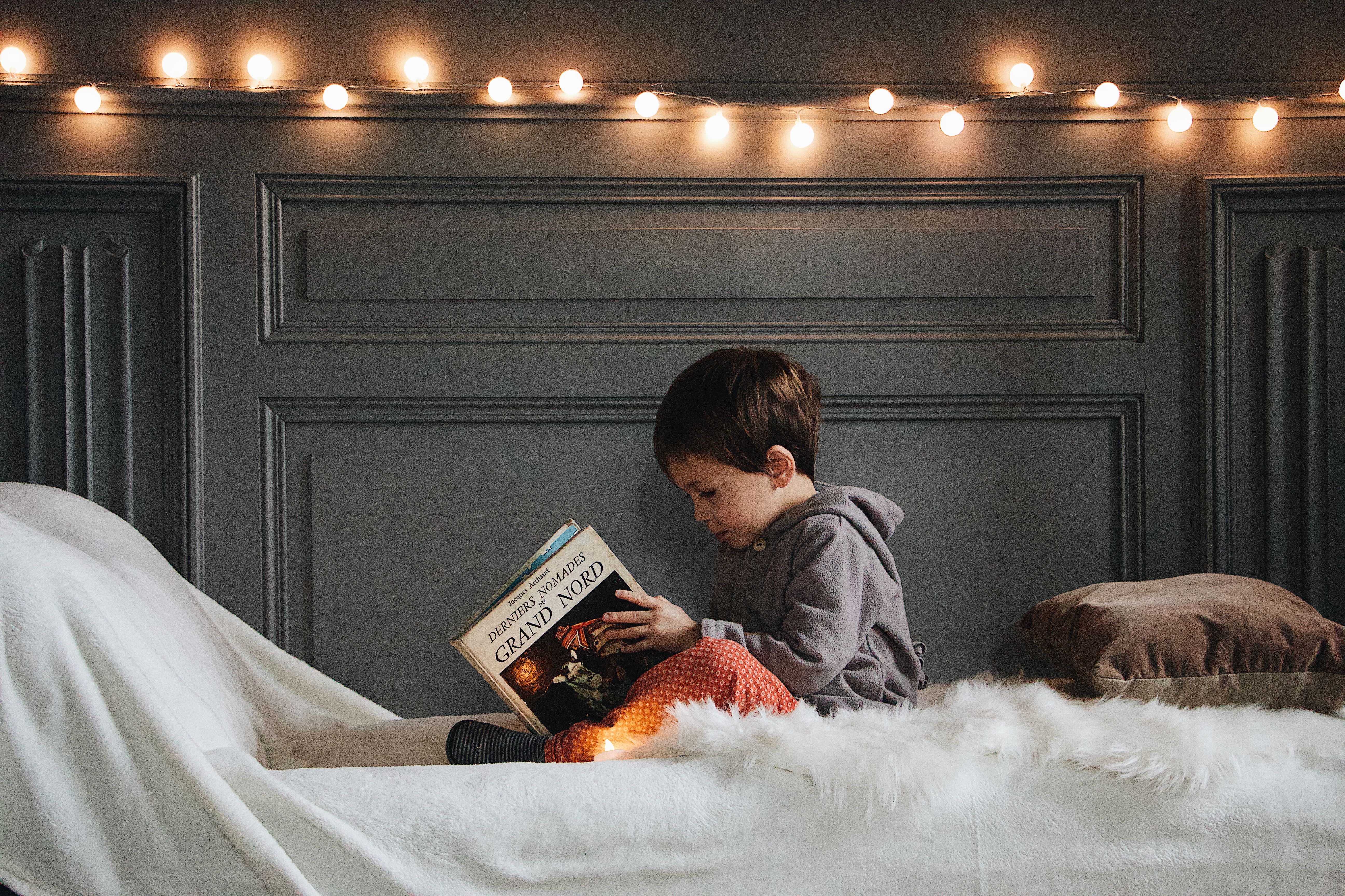 A man with dark curly hair, beard, and glasses sitting on a gray sofa reading a red book while a young boy leans affectionately on his shoulder, both smiling warmly.