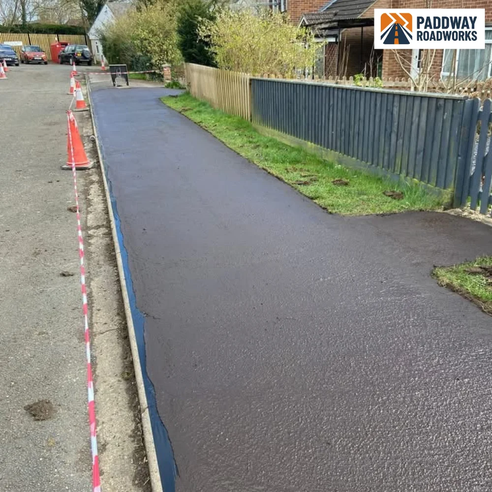 Long red/maroon-asphalt strip resurfaced between houses, blue safety barriers.