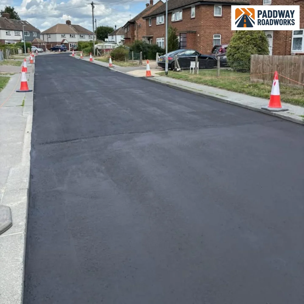 Wide road being newly tarmac-resurfaced between residential houses.