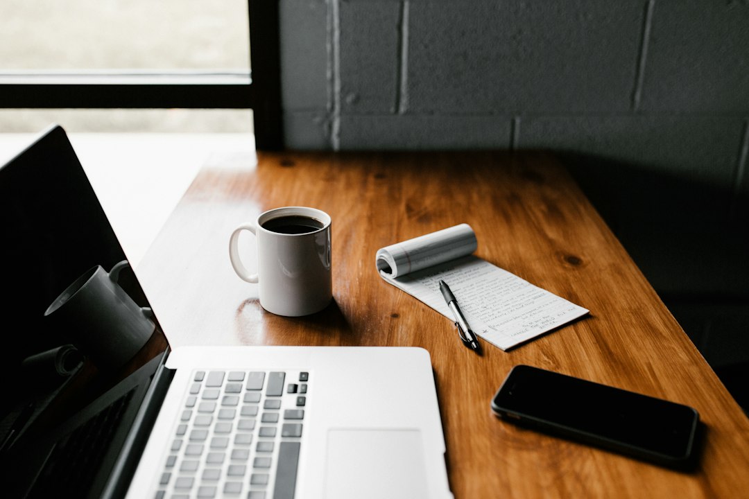 Laptop, white coffee mug, notepad with pen and a phone on a wooden desk beside a window and grey brick wall.