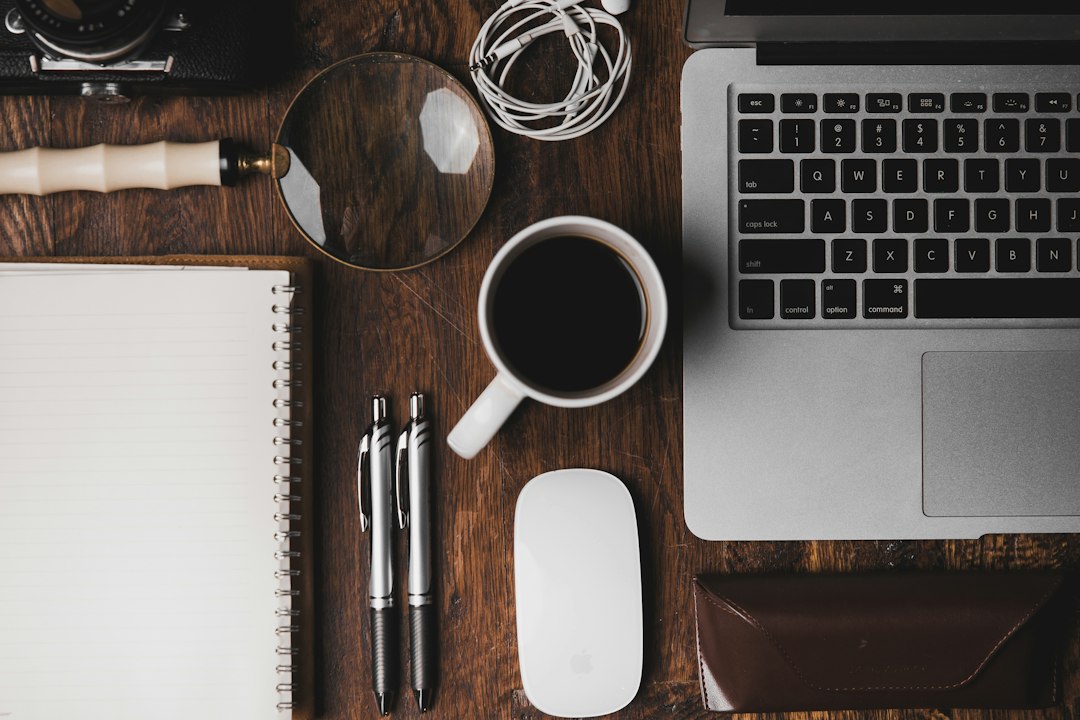Overhead view of a wooden desk with laptop, coffee cup, mouse, pens, notebook, magnifying glass and earphones.