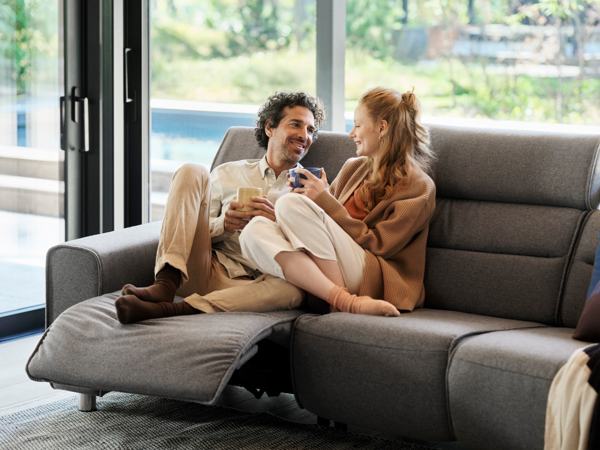 A couple sitting together on a grey Stressless sofa with the footrest extended, beside a garden window