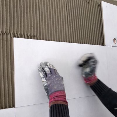 Close-up of gloved hands placing large white wall tiles on a mortar base