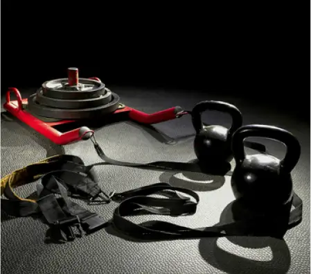 Kettlebells, weight sled, straps, and gloves on a gym floor under dim lighting.