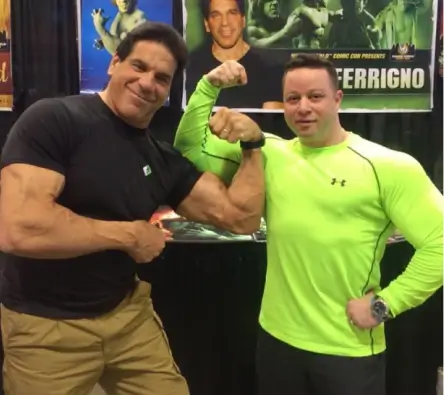 Jamie Mushlin, owner of Red Fitness Lounge, poses flexing his muscles alongside a client at an event. Jamie dons a black shirt while his client sports a bright green one. Posters are visible in the background.