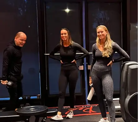 Three individuals wearing athletic clothing stand and smile in a gym; two women in the foreground and a man holding a dumbbell in the background.