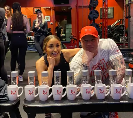 Two people posing behind a row of Red-branded mugs and water bottles in a gym. The woman is smiling and leaning on her hands, while the man rests his chin on his hand. Others exercise in the background.