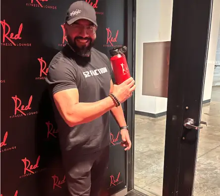 Man in a black t-shirt and cap smiling and holding a red water bottle in front of a door with a "Red Fitness Lounge" backdrop.