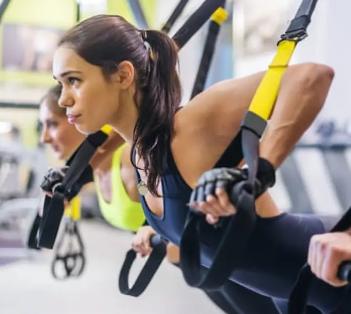 Three people exercise using TRX suspension trainers at the Red Fitness Lounge Personal Training center. They are focused, wearing athletic clothing and gloves, and are performing a push-up movement.