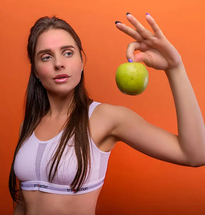 A woman wearing a sports bra holds a green apple with her right hand against an orange background, embodying the vibrant energy of the Red Fitness Lounge.