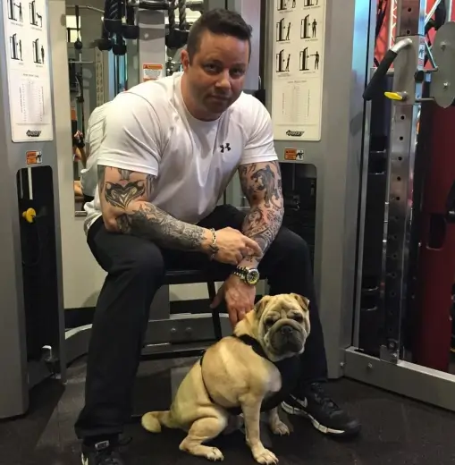 Jamie Mushlin, owner of Red Fitness Lounge training center, sits on gym equipment in a white t-shirt and black pants, showing off his arm tattoos. Beside him, a bulldog on a leash sits attentively.