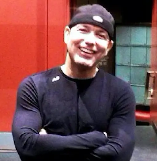 Joey Neal, a certified personal trainer and certified Yoga instructor with short dark hair and wearing a black jacket, stands with arms crossed against the vibrant red backdrop of Red Fitness Lounge.