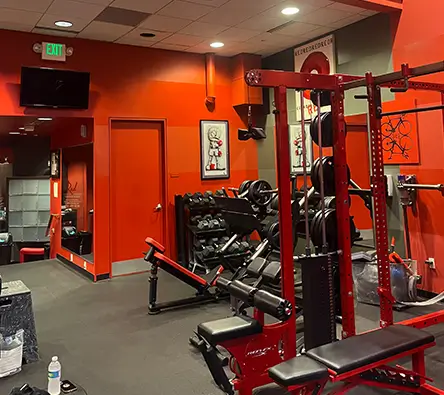 An indoor gym with various weights and benches, a mirror on the wall, and an automated external defibrillator (AED) unit mounted beside the mirror.