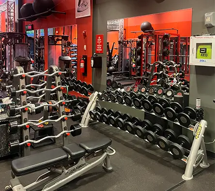 An indoor gym with various weights and benches, a mirror on the wall, and an automated external defibrillator (AED) unit mounted beside the mirror.