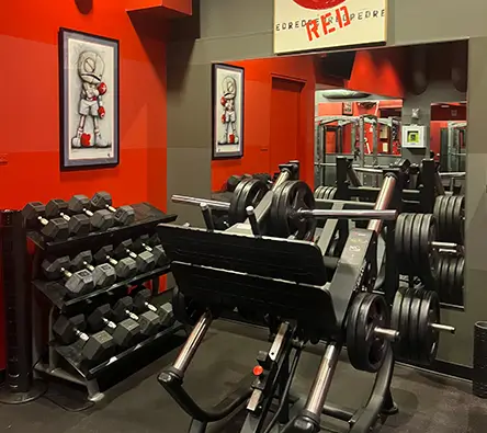 A gym room with a mirrored wall, dumbbell rack, and a leg press machine loaded with weight plates. Red and black decor with framed sports-themed artwork on the walls.