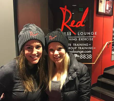 Two smiling women wearing matching black beanies and jackets stand in front of a door with the logo "Red Fitness Lounge" and contact information displayed.