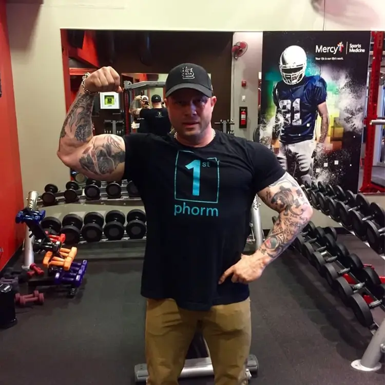 A man in a gym flexes his right arm while wearing a black cap and a black t-shirt with "1st phorm" written on it. Behind him, various gym equipment and a poster featuring a football player are visible. This is Jamie Mushlin, owner of Red Fitness Lounge.