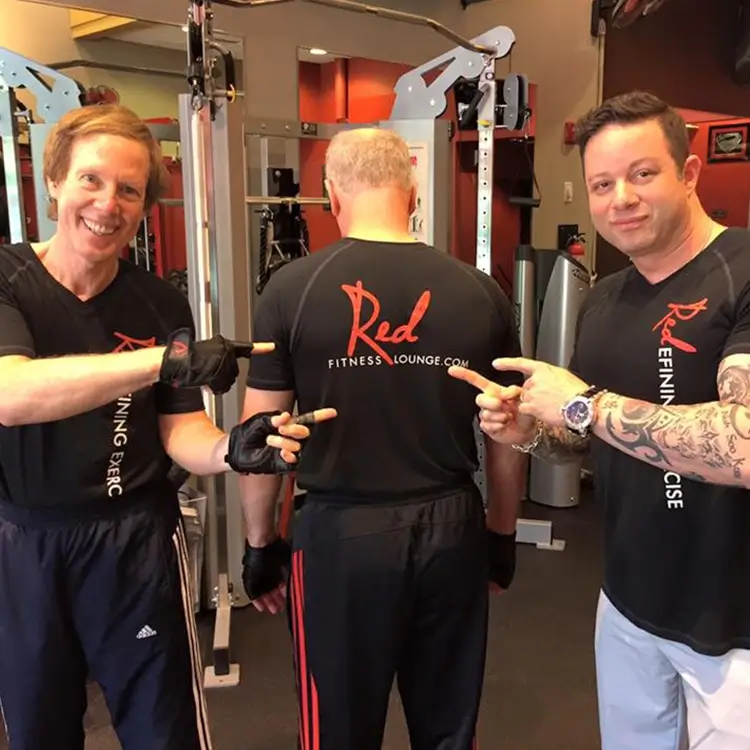 Three men at a gym, with two in the foreground pointing at the back of the third man's shirt that reads “Red Fitness Lounge.” Gym equipment is visible in the background.