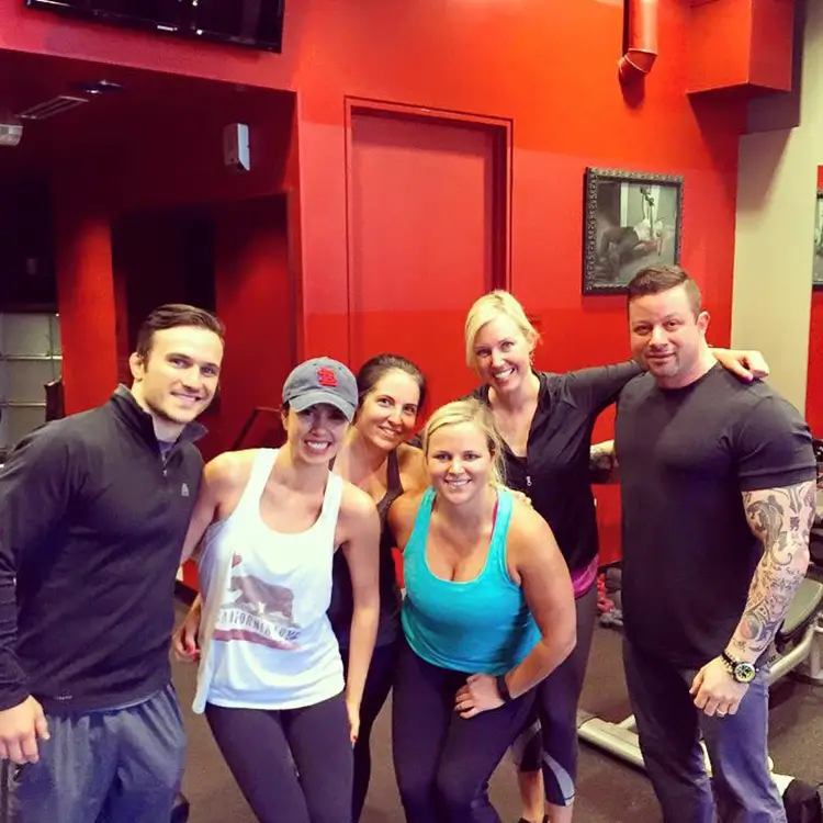 Six people posing together in a gym, smiling. Two men and four women in fitness attire stand arm in arm.