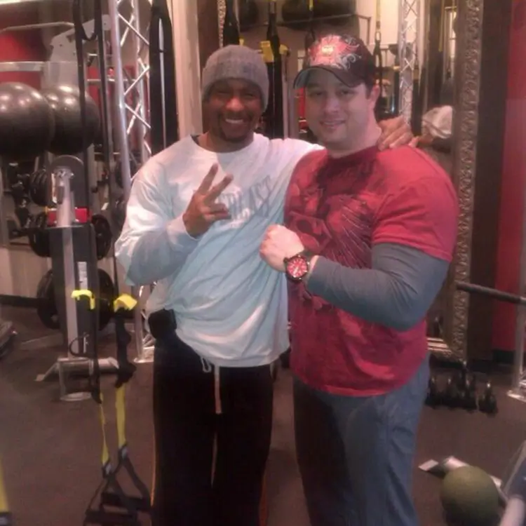 Two people are posing for a photo in a gym, with exercise equipment in the background. One is making a peace sign while the other is smiling and placing an arm around the first person's shoulder.