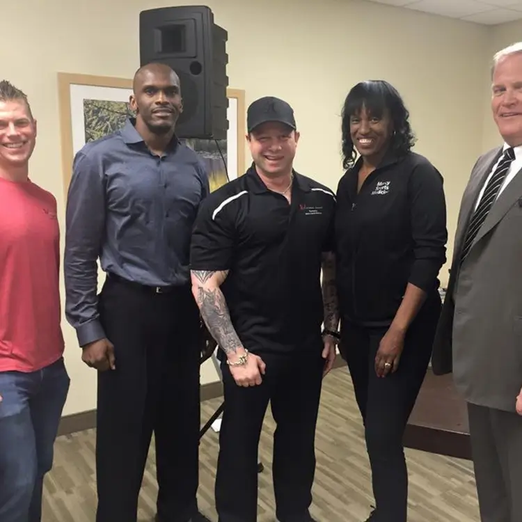 Five people standing indoors; on the left, a man in a red shirt; next, a man in a blue shirt; center, a man in black gym attire; next, a woman in black attire; and on the right, a man in a suit.