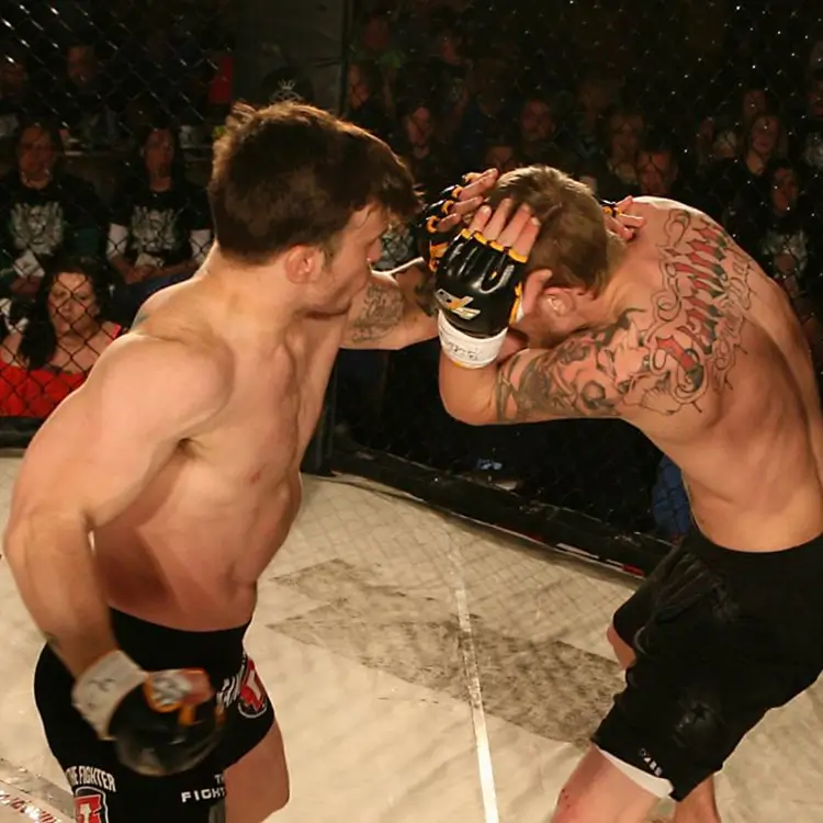 Two mixed martial arts fighters in a cage fight, with one throwing a left punch and the other guarding his head with both hands. Both are shirtless and wearing black shorts and gloves.