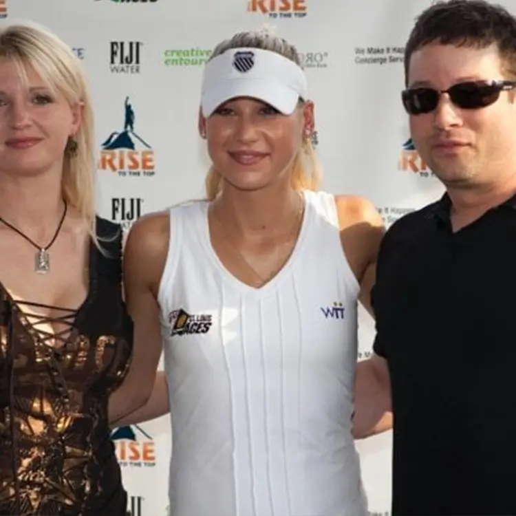 Three individuals posing at a sports event; a woman in a gold top, a woman in a white sports outfit and cap, and a man in dark sunglasses and a black shirt. Event banners are visible in the background.