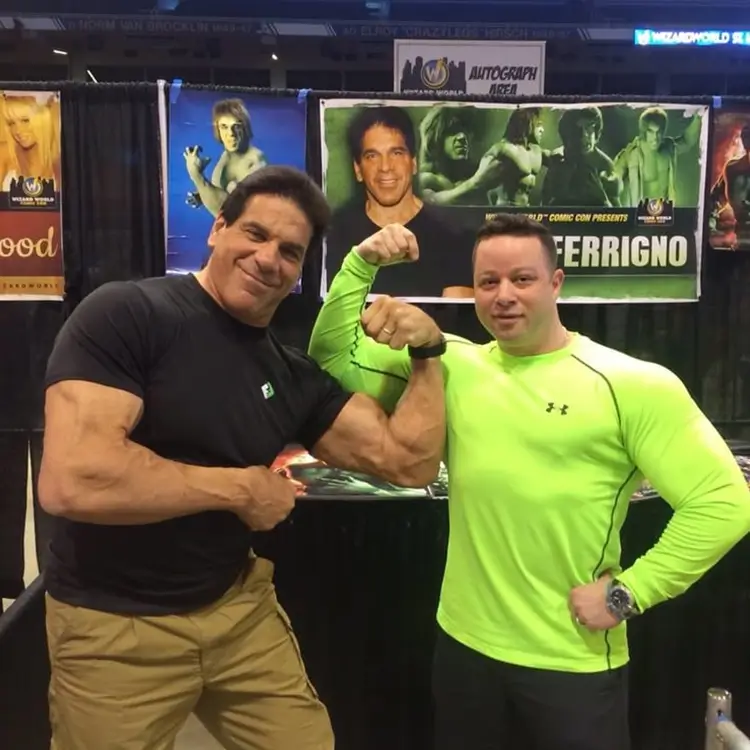 Jamie Mushlin, owner of Red Fitness Lounge, poses flexing his muscles alongside a client at an event. Jamie dons a black shirt while his client sports a bright green one. Posters are visible in the background.