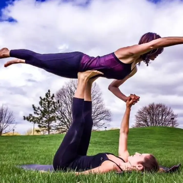 Two individuals perform acro yoga on a grassy field under a partly cloudy sky. The base person lies on their back, holding the flyer in a plank position elevated on their feet.