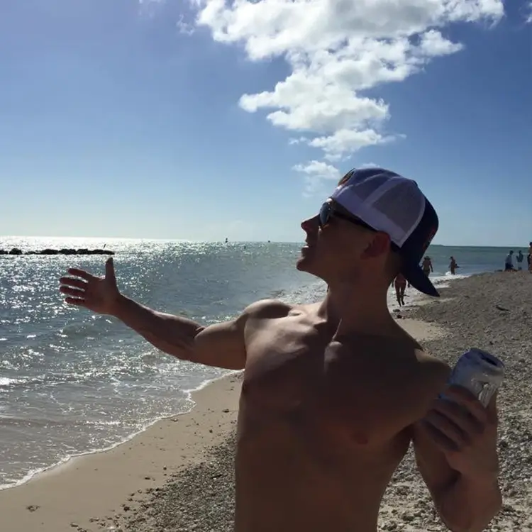 A shirtless person wearing a backwards cap and sunglasses stands on a beach holding a drink, with one arm outstretched toward the ocean on a sunny day.