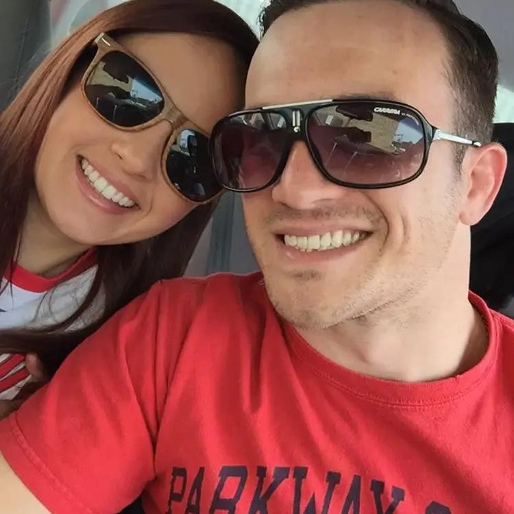 A man and a woman in sunglasses smile while taking a selfie inside a car.