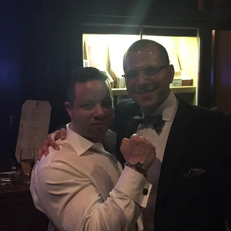 Two men posing together indoors, one with his arm around the other. The man on the right is wearing a suit and bow tie; the man on the left, Jamie Mushlin, the owner of Red Fitness Lounge, is wearing a white shirt. They are in a dimly lit environment.