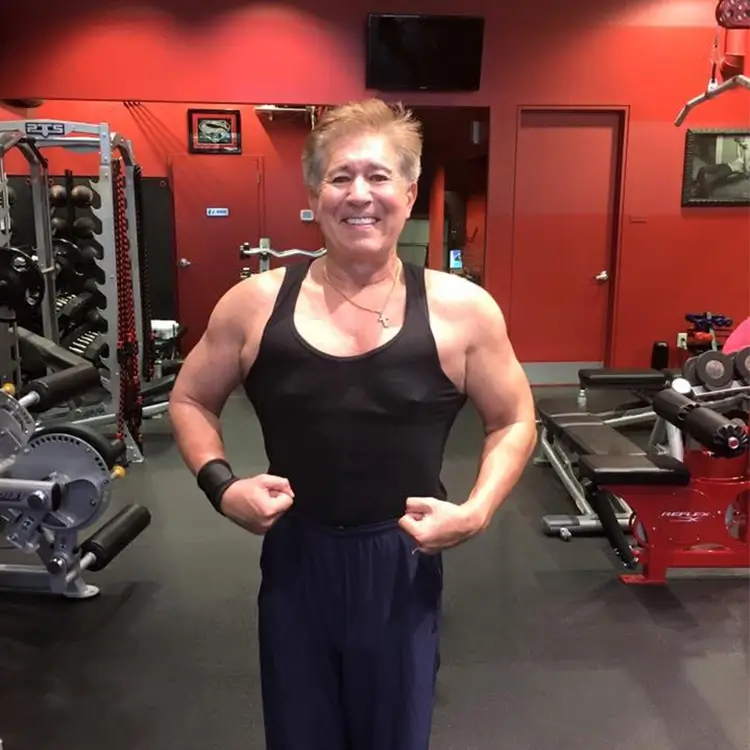 Person in a black tank top and navy pants posing and flexing muscles in a gym setting with various workout equipment.