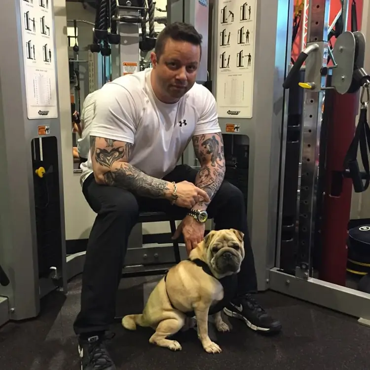 Jamie Mushlin, owner of Red Fitness Lounge training center, sits on gym equipment in a white t-shirt and black pants, showing off his arm tattoos. Beside him, a bulldog on a leash sits attentively.