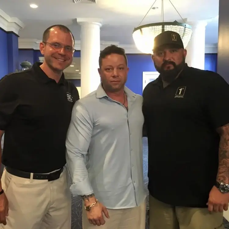 Three men are standing together indoors with one man in a light blue shirt and the other two in dark shirts. They are posing for the camera and appear to be in a well-lit room with blue walls.