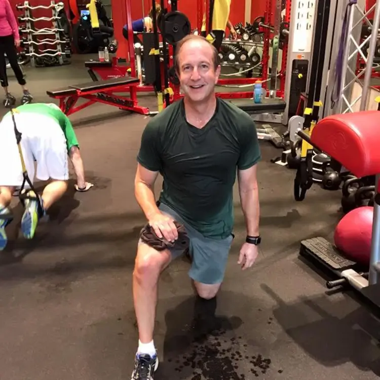 A man at the gym is doing lunges, wearing a green shirt and shorts. Exercise equipment is visible in the background, and another person is exercising nearby.